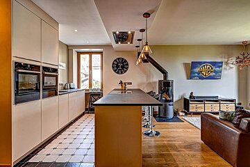 Modern open-plan kitchen/living room with integrated appliances, island, wood stove, TV, and contrasting tiled/wood flooring.