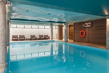 An indoor pool with bright blue water, a wavy reflective teal ceiling, lounge chairs, and a lifebuoy on a wooden wall.