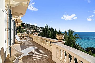 Elegant stone balcony with white sun loungers overlooking a stunning blue sea, green hills, and coastal trees.