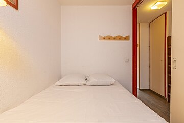 A sparse bedroom with a bed, white linen, and two pillows. White textured walls feature a wooden coat rack. A red-framed doorway leads to a bright hallway.