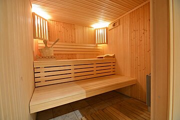 A bright, modern wooden sauna interior with tiered slatted benches, warm backlighting, and a traditional wooden bucket with a ladle.