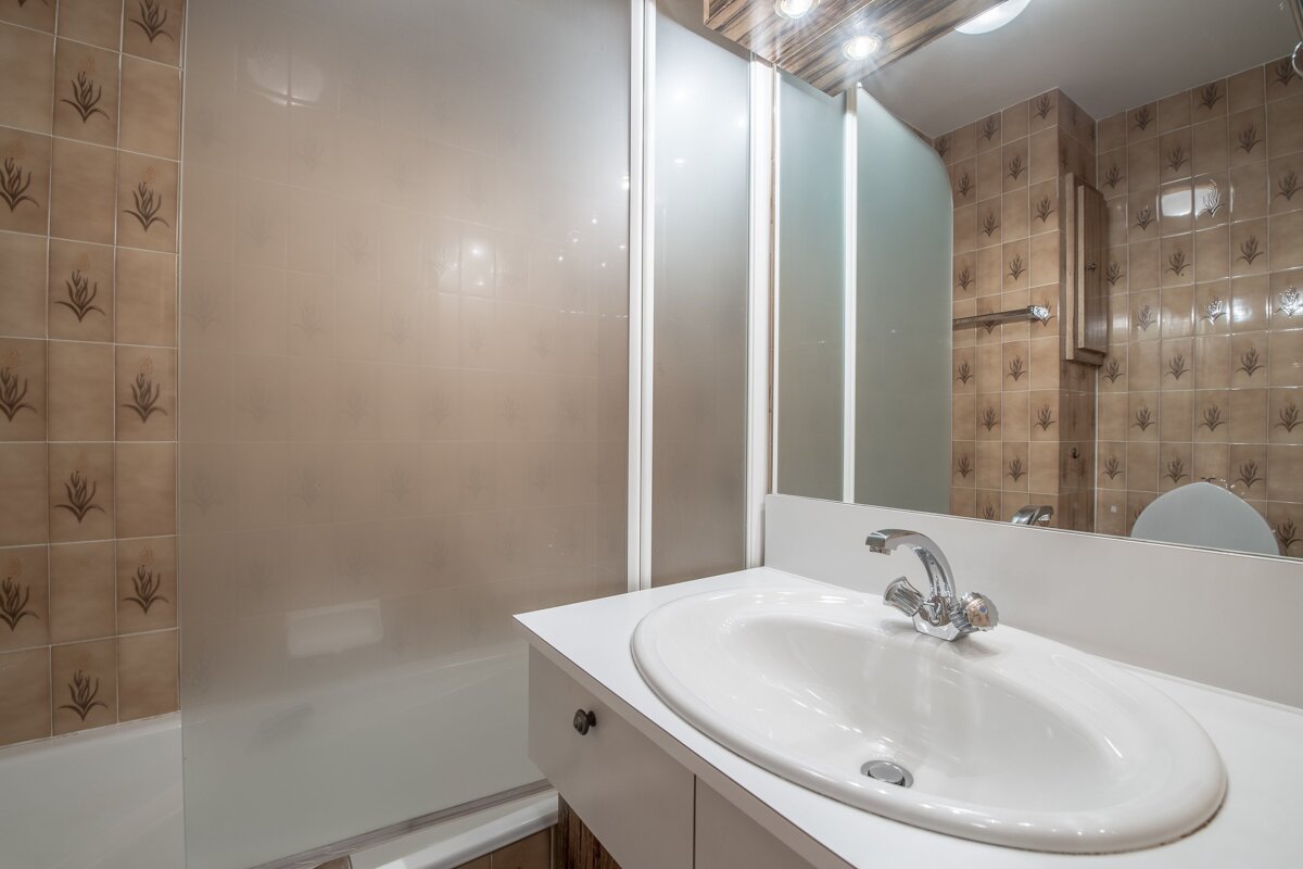 A bathroom with patterned brown tiles, a white vanity & sink, and a frosted glass shower. A mirror reflects the toilet.
