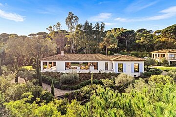 A modern Mediterranean villa with a terracotta tiled roof, surrounded by lush green trees and gardens under a clear blue sky.