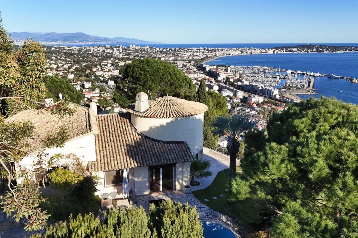 An aerial view reveals a charming house on a verdant hill, overlooking a vibrant coastal city, a bustling marina, and the sparkling blue sea.