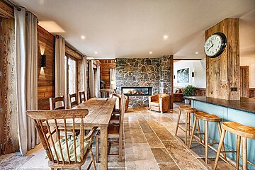 A rustic open-plan room with a long wooden dining table, a stone fireplace, wood-paneled walls, and a kitchen island with bar stools.