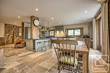 A spacious, rustic open-plan kitchen and dining room featuring stone floors, wooden beams, sage green cabinetry, a large island, and a cozy armchair.