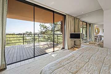 A bright, modern bedroom featuring large sliding glass doors that open onto a wooden deck balcony with a scenic tree-filled view.