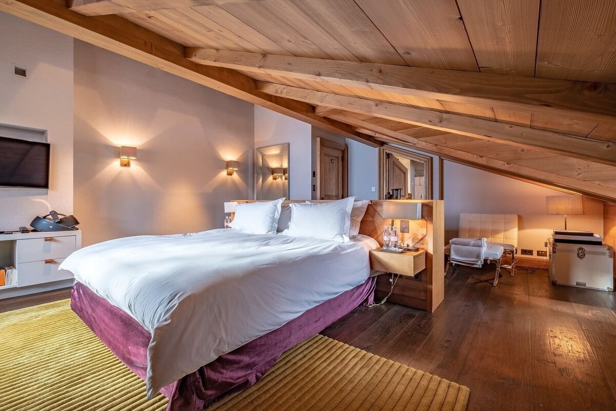 A modern, luxurious attic bedroom featuring a large white bed, warm ambient lighting, and a prominent sloped wooden ceiling with exposed beams.