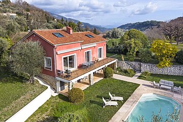 A red house with a terraced balcony and a swimming pool, surrounded by a green lawn and trees, nestled in rolling hills under a partly cloudy sky.