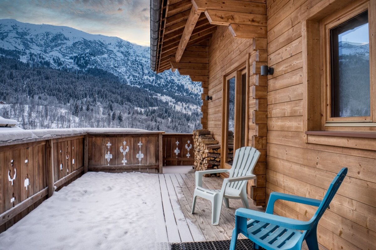 A snowy wooden chalet balcony with two chairs and stacked firewood, facing a majestic view of snow-covered mountains and evergreen trees under a cloudy sky.
