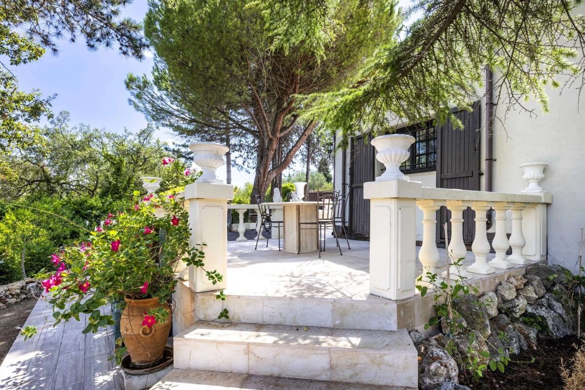 A charming outdoor stone patio with steps, a balustrade, and a dining set under a large tree. Lush greenery and a white house with shutters are visible.
