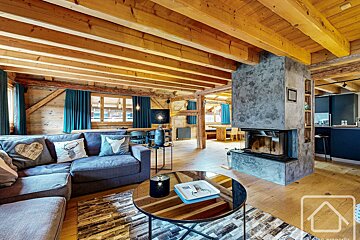 Rustic, open-plan living room with exposed wooden beams, a gray sectional sofa, modern concrete fireplace, and teal curtains.