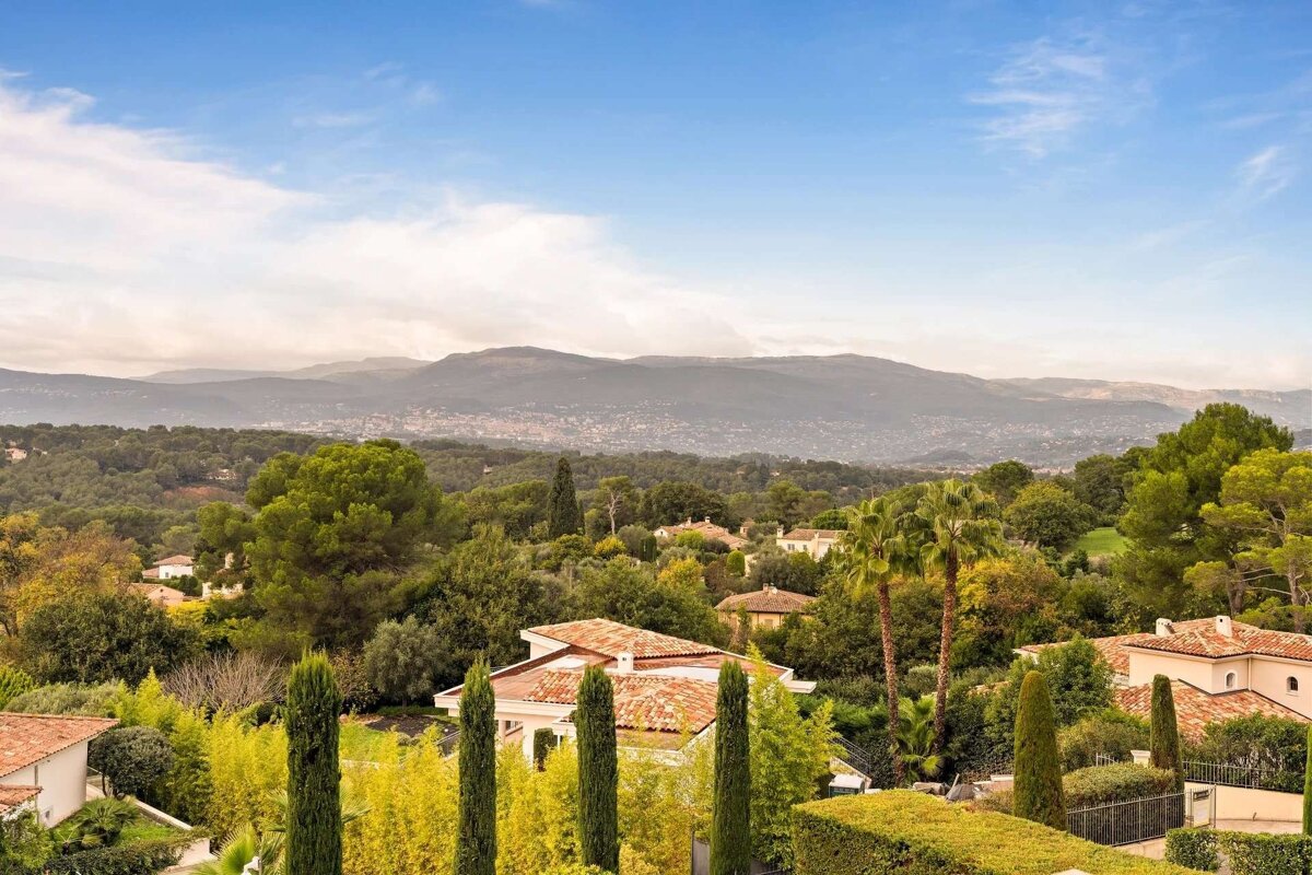 Panoramic view of a lush green landscape with red-tiled houses nestled among trees. Distant mountains and a city are visible under a partly cloudy blue sky.