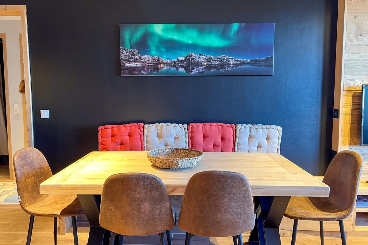 A stylish dining area with a light wood table, brown chairs, and a cushioned bench. A vibrant Aurora Borealis artwork hangs on the dark wall.