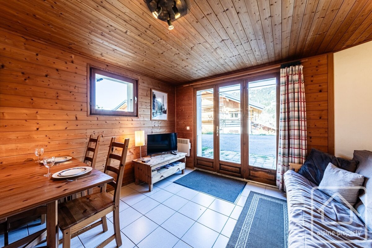 A cozy, wood-paneled room with a dining table, sofa, TV, and glass doors opening to an outdoor view. Features a light-tiled floor and plaid curtains.