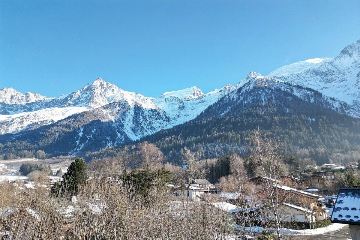 Snow-capped mountain peaks towering over a small valley village under a clear blue sky.