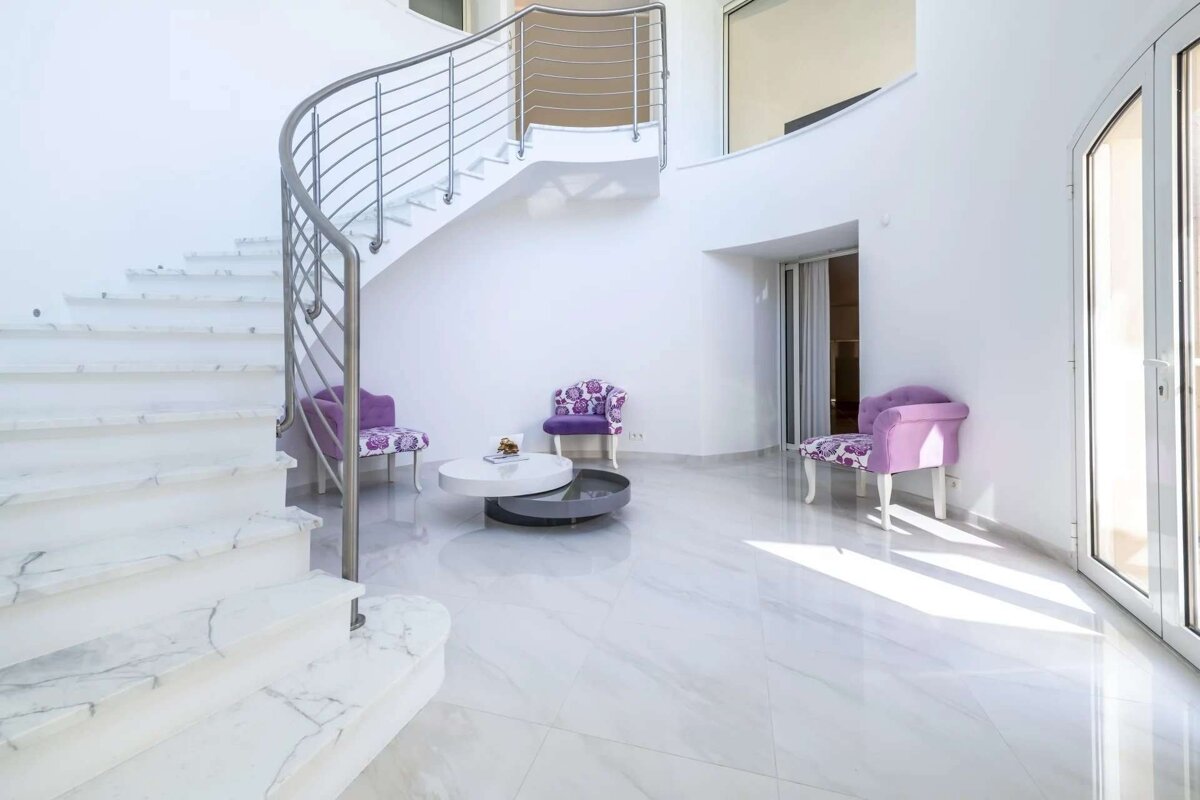A bright, modern white interior featuring a curved marble staircase with a steel railing, three purple armchairs, and a round coffee table.