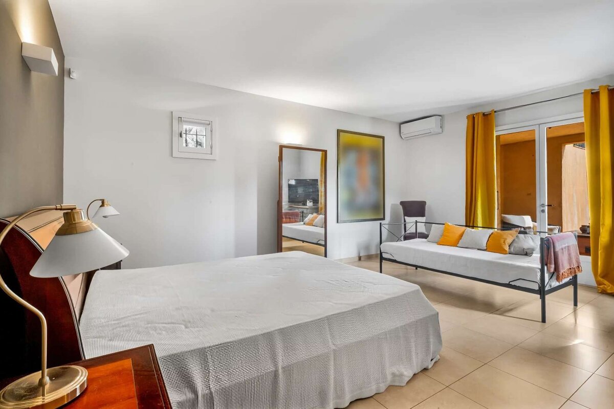 A bright bedroom with a large bed, daybed with colorful cushions, yellow curtains, and a sliding glass door leading to an outdoor area.