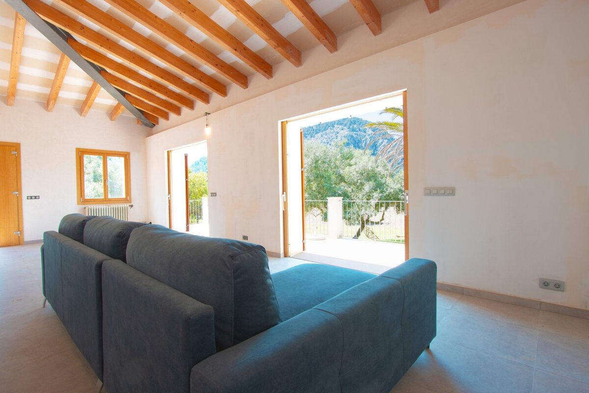 A bright living room with a gray sofa and exposed wooden beams. Large glass doors open to a sunny patio with trees and mountains in the distance.