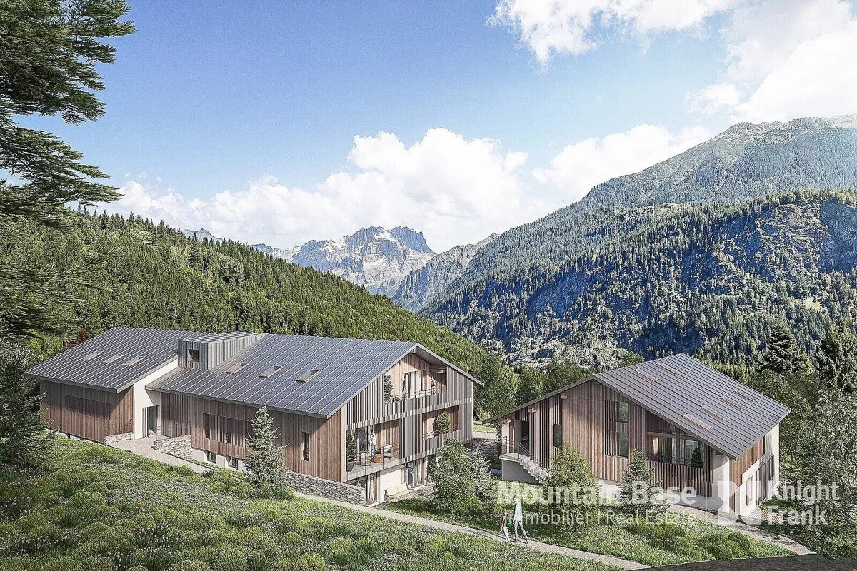 Two modern, wood-clad mountain chalets nestled in a lush, forested valley under a clear blue sky, with towering peaks in the background.