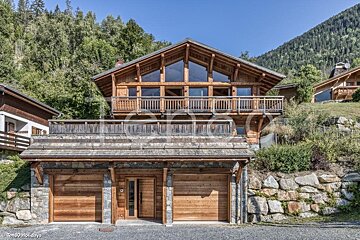 A beautiful multi-level wooden cabin with a stone-accented garage, surrounded by lush green trees on a mountain slope.