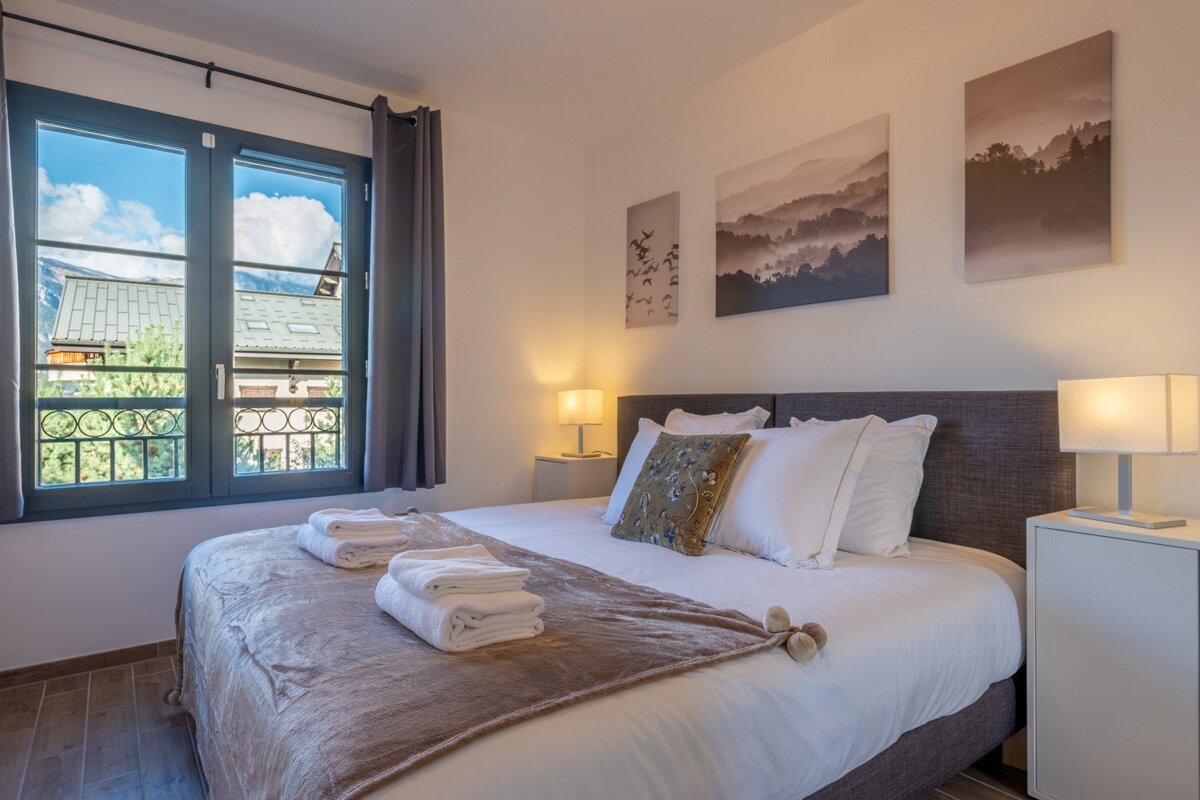A cozy bedroom with a large bed, white linens, bedside lamps, mountain artwork, and a window revealing a view of buildings and distant mountains.