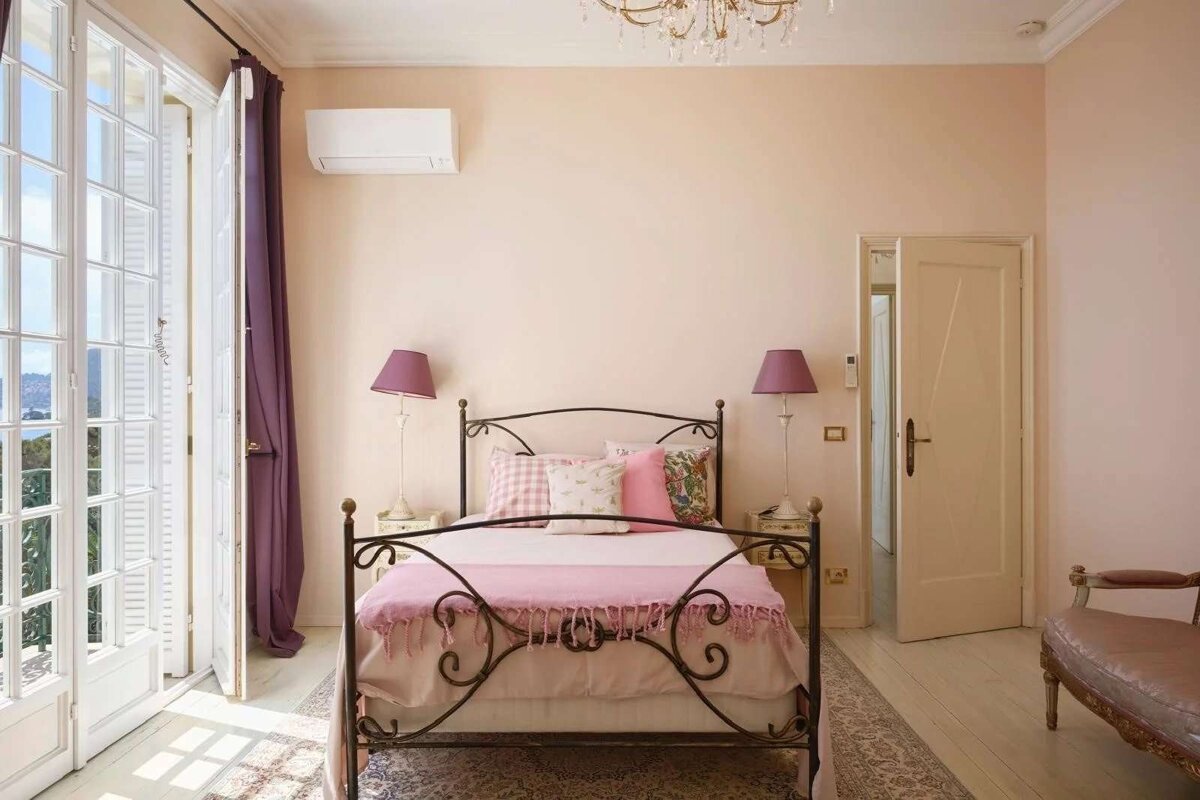 A romantic bedroom with a wrought iron bed, pink linens, and open French doors leading to a scenic balcony. Soft lighting and pastel walls create a serene space.