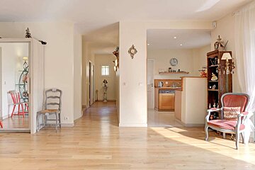 A sunlit, open home interior with light wood floors, leading to a kitchen and a hallway. Features include decorative chairs and ornate wall decor.
