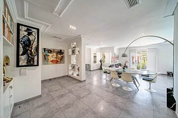 A spacious, modern dining and living room with grey tiled floors, white walls, colorful art pieces, and a glass-top table.