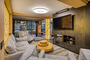 A contemporary living room featuring gray sofas, a tree stump coffee table, a large TV, and striking backlit amber walls, opening to a patio.