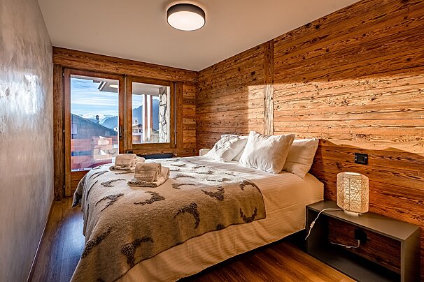 A cozy, rustic bedroom featuring warm wood-paneled walls, a large bed, and a glass door opening to a mountain view.