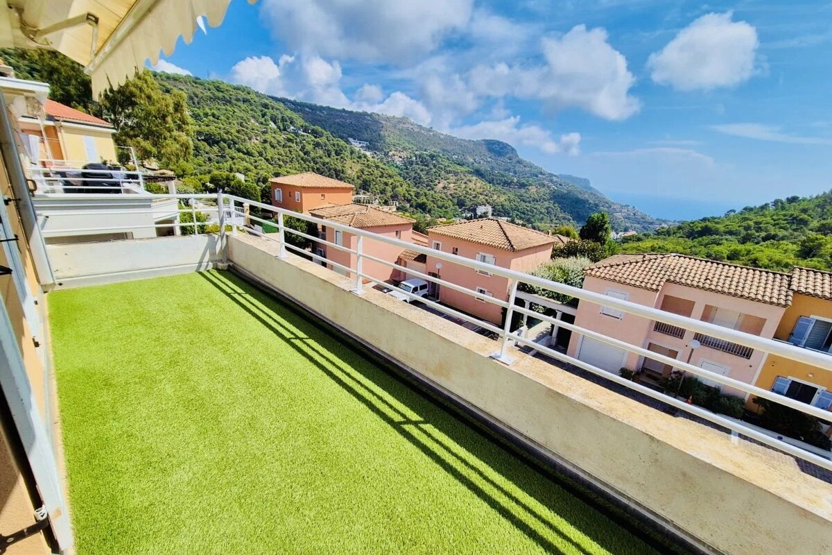 Balcony with artificial grass overlooking Mediterranean-style houses nestled in green hills under a bright blue sky.