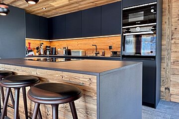 Modern kitchen featuring a rustic wood-paneled island with bar stools, sleek black cabinetry, and warm wood-paneled walls and ceiling.