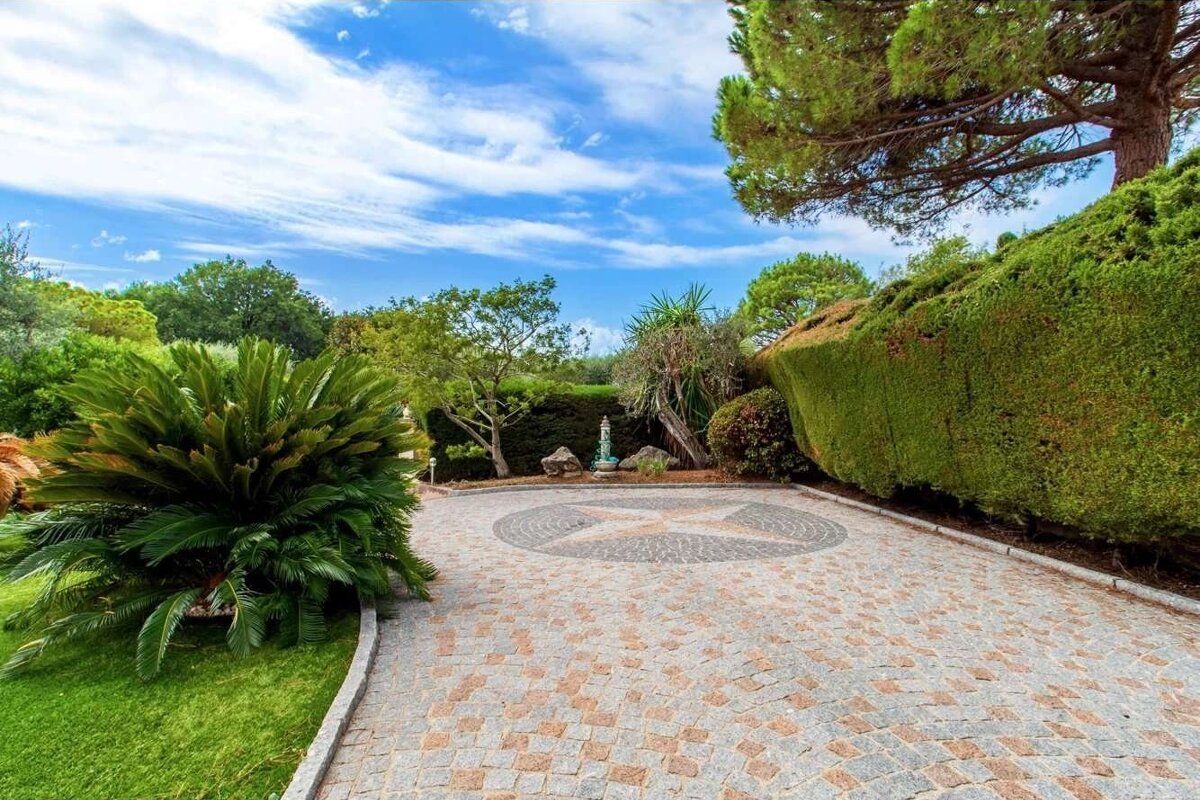 A cobblestone driveway with a star design leads through a lush garden with diverse green foliage, large trees, and a small fountain under a bright blue sky.