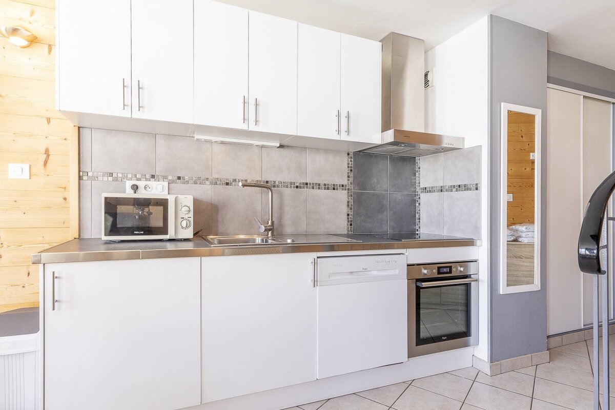 A modern kitchen with white cabinets, a stainless steel counter, microwave, oven, dishwasher, tiled backsplash, and a wooden wall.