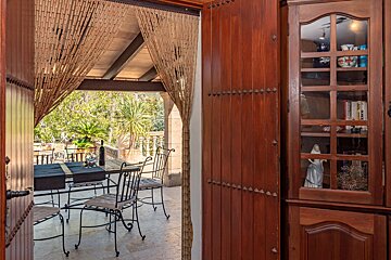 A view from indoors through a bamboo curtain to an outdoor patio with a dining table, wine, and lush garden. A wooden cabinet is on the right.