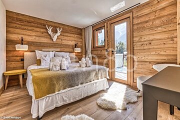 Cozy rustic chalet bedroom with wood-paneled walls, a plush bed, faux deer skull decor, furry rugs, and a glass door to the outside.