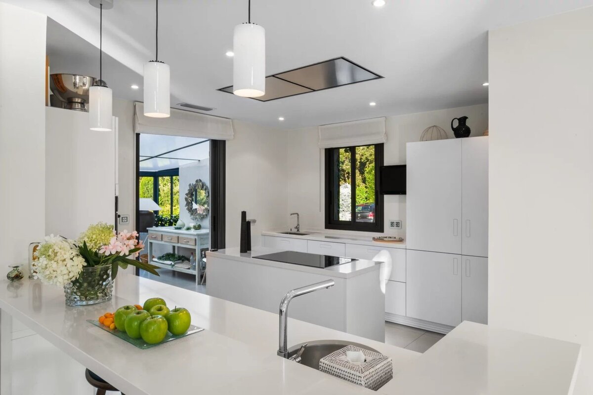 A sleek white modern kitchen featuring a double island, glass pendant lights, a bowl of green apples, and a floral arrangement.
