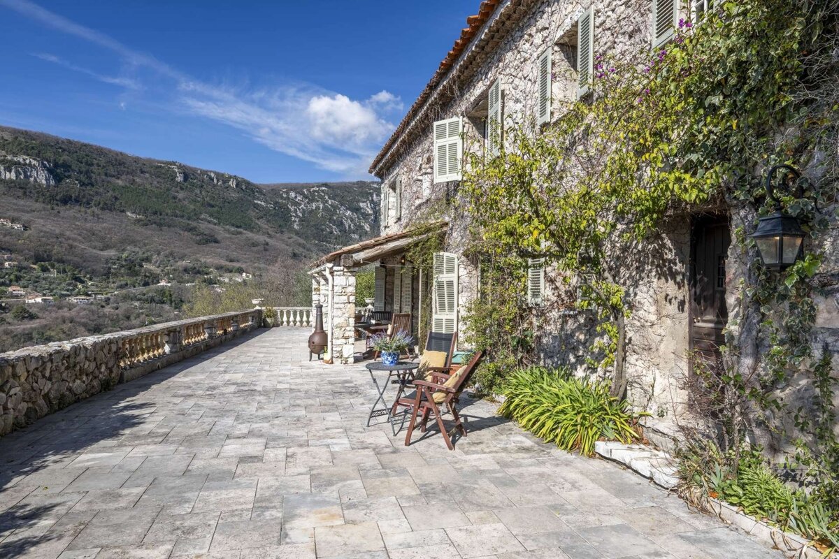 A scenic stone house with a spacious paved patio overlooking mountains under a blue sky. Vines adorn the walls, with outdoor seating.
