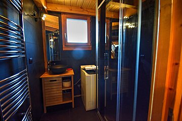 A modern bathroom with dark tiles, wooden ceiling, shower, wooden vanity with black basin, washing machine, and chrome towel warmer.