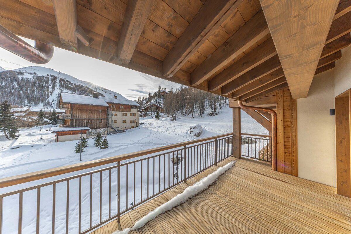 A wooden balcony looks out over a snowy alpine landscape with traditional ski chalets and snow-covered mountains.