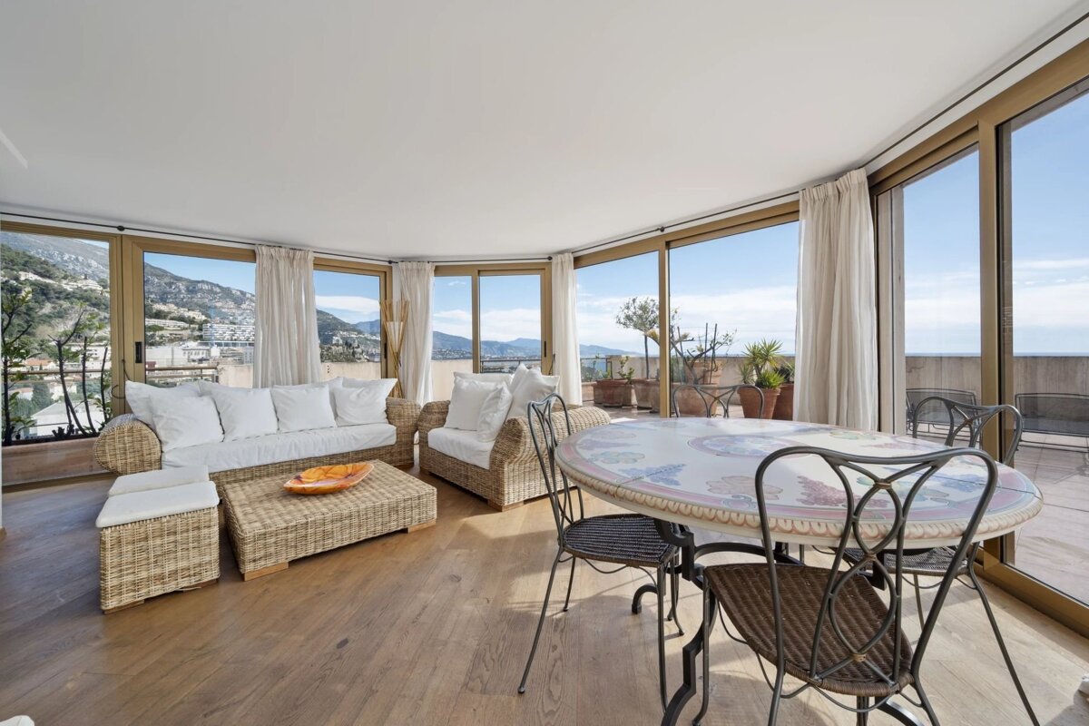 A sunlit patio room featuring wicker sofas, a dining table set, and large windows with panoramic coastal views.