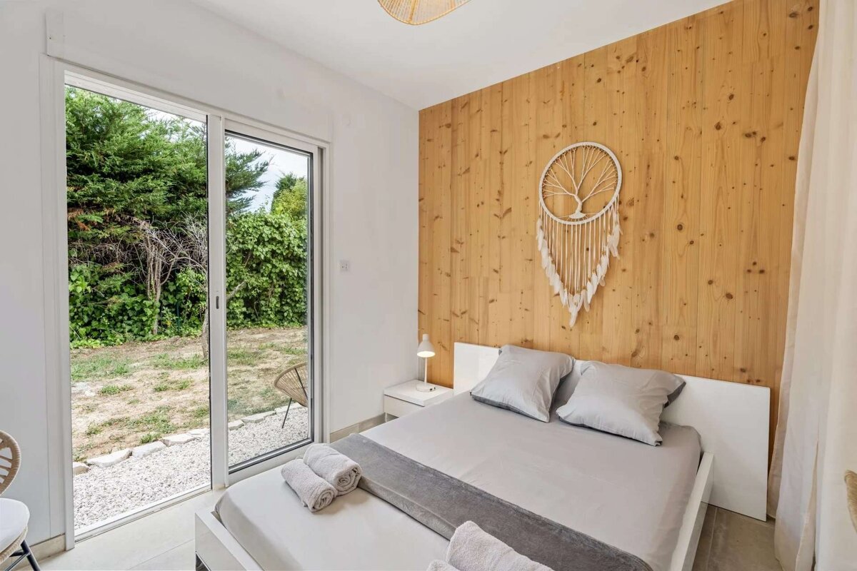 Bright bedroom featuring a bed, wood paneled wall with a Tree of Life dreamcatcher, and a glass door leading to a green garden.