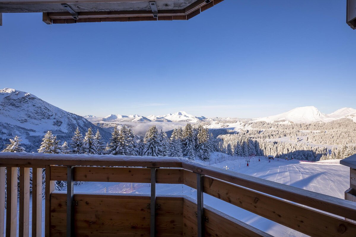 View from a balcony of snow-covered mountains, frosted pine trees, and a sunny ski slope beneath a clear blue sky.