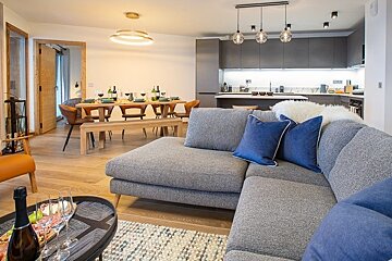 Cozy open-plan living room with a grey sectional, blue cushions, wooden dining table, and a modern kitchen in the background.