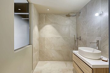 A modern bathroom with light-colored marble-look tiles, featuring a walk-in shower and a sleek wooden vanity with a white vessel sink.