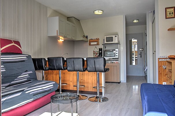 A small studio apartment featuring a living area with sofas, a wooden bar with black stools, and a compact kitchen in the background.