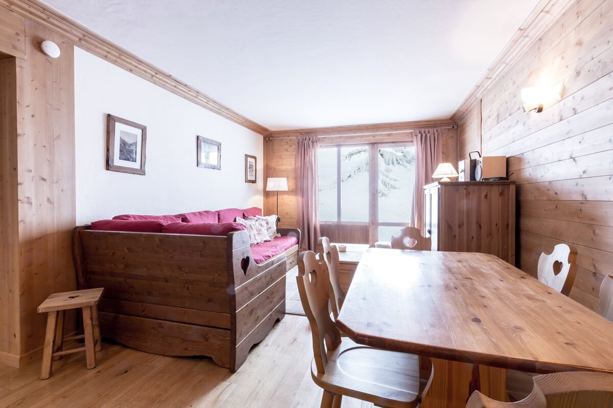 Rustic wooden chalet interior featuring a dining table, a cozy red sofa, and a snowy view through the window.
