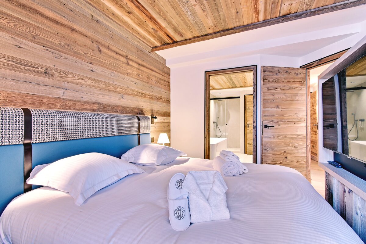 Modern bedroom with wooden walls and ceiling, a blue and patterned headboard, and white bedding. Slippers and towels are on the bed, a bathroom is visible.