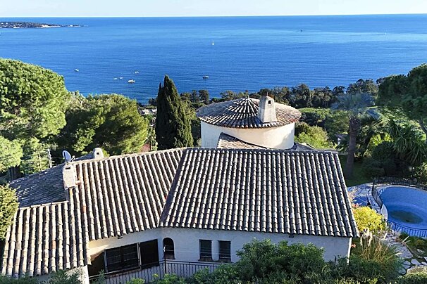 Aerial view of a Mediterranean house with tiled roofs, a round tower, and a blue pool, surrounded by trees, overlooking a deep blue sea with boats.